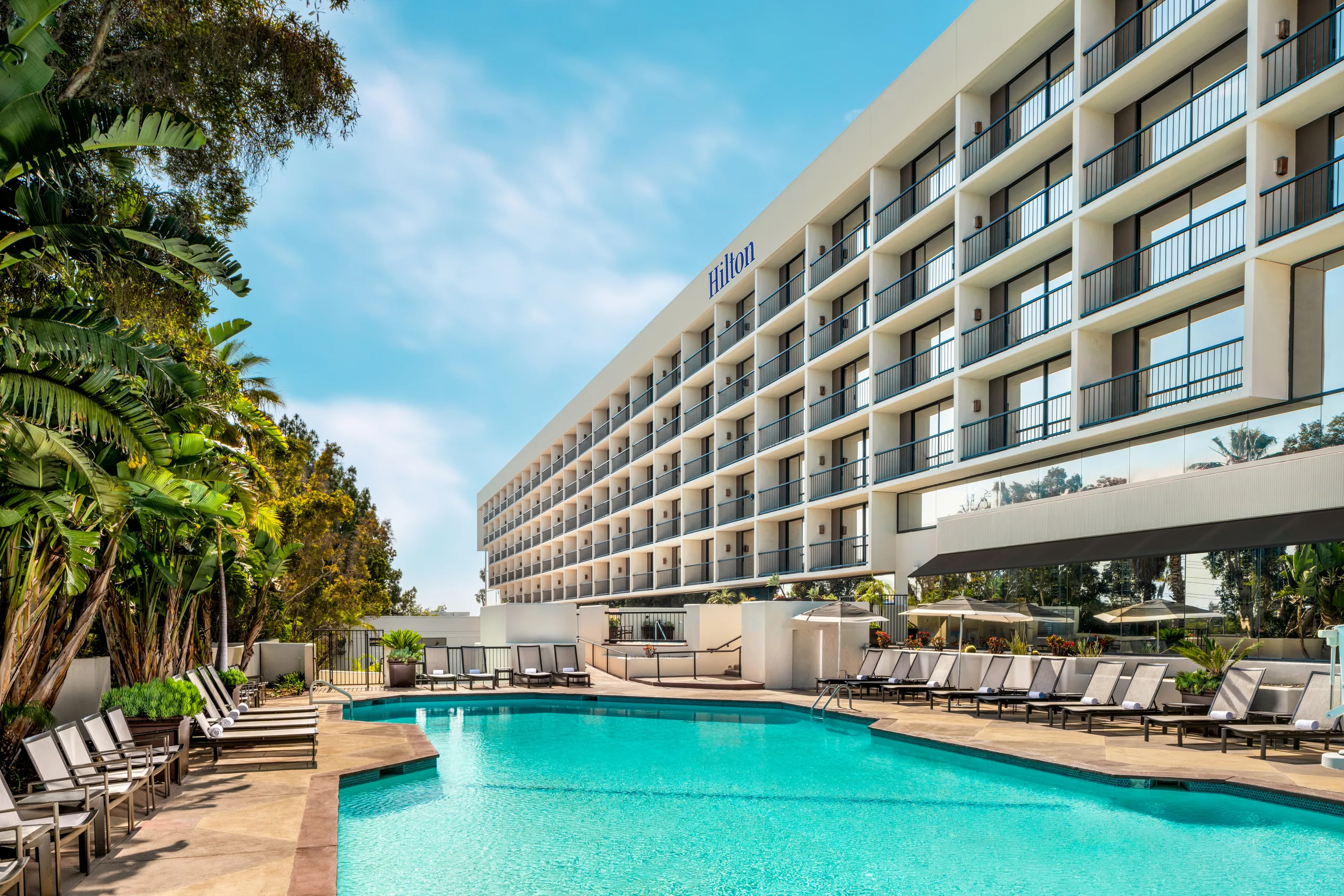 A large outdoor swimming pool surrounded by lounge chairs sits in front of the multi-story Hilton Orange County Costa Mesa hotel with balconies, shaded by palm trees and set against a backdrop of blue sky.