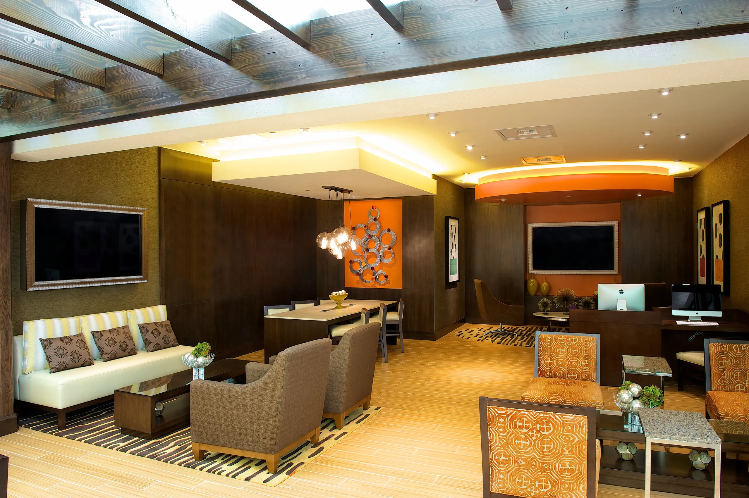 Modern lounge at Hilton Orange County Costa Mesa with contemporary furniture—beige and brown chairs, a white sofa, desks with computers, decorative lighting, and orange accents. Wooden beams cross the ceiling and art decorates the walls.