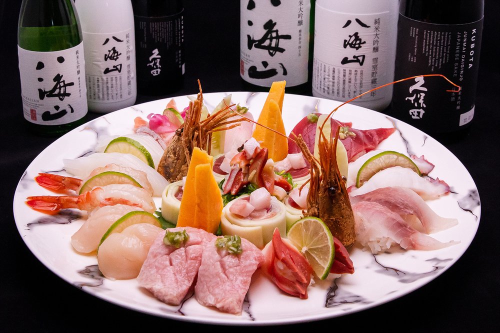 A marble plate with assorted sushi and sashimi, including shrimp, tuna, salmon, and garnishes. Bottles of sake are arranged in the background against a black backdrop.