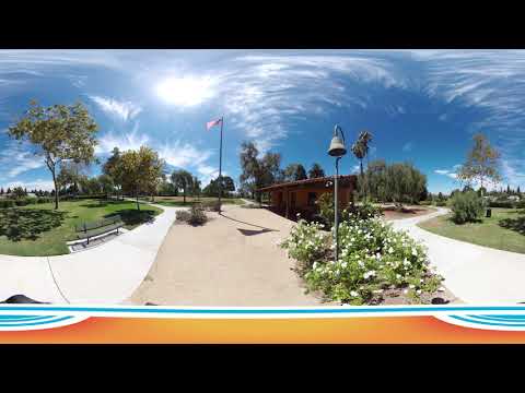 A 360-degree outdoor view of Estancia Park Costa Mesa with green lawns, trees, flowering bushes, a flagpole with the American flag, and a brown building under a bright blue sky with wispy clouds.