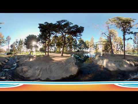 A 360-degree view of TeWinkle Park Costa Mesa showcases tall trees, a walking path, a small stream, sunlight filtering through the branches, and blue sky, all framed by a colorful orange and blue border at the bottom.