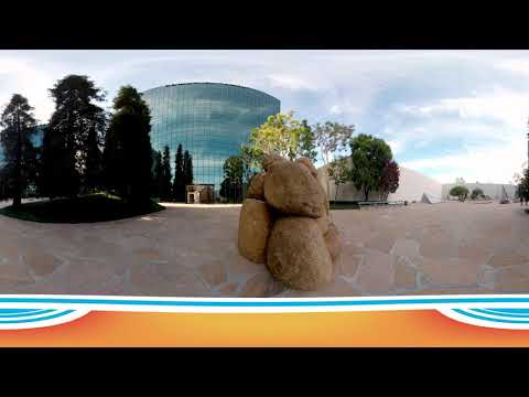 A group of large boulders is stacked in a modern outdoor plaza at the Noguchi Garden Costa Mesa, with stone pavement, trees, and a glass-fronted building under a blue sky. A curved, colorful graphic borders the bottom of the image.