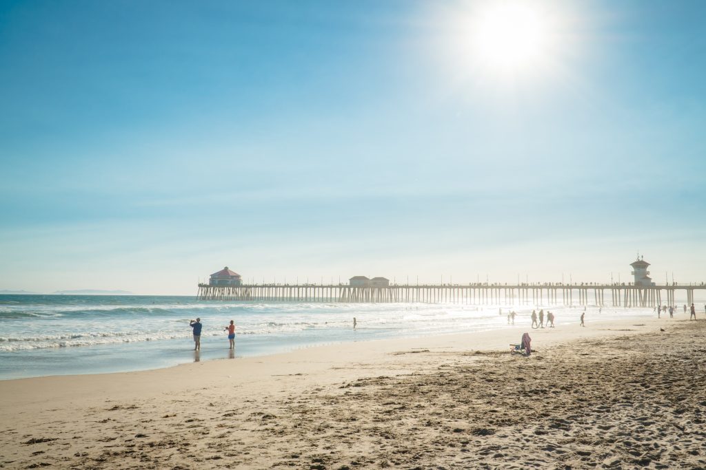 People walk and relax on a sandy beach near gentle waves, with a long pier and buildings extending into the ocean under a bright sunny sky.