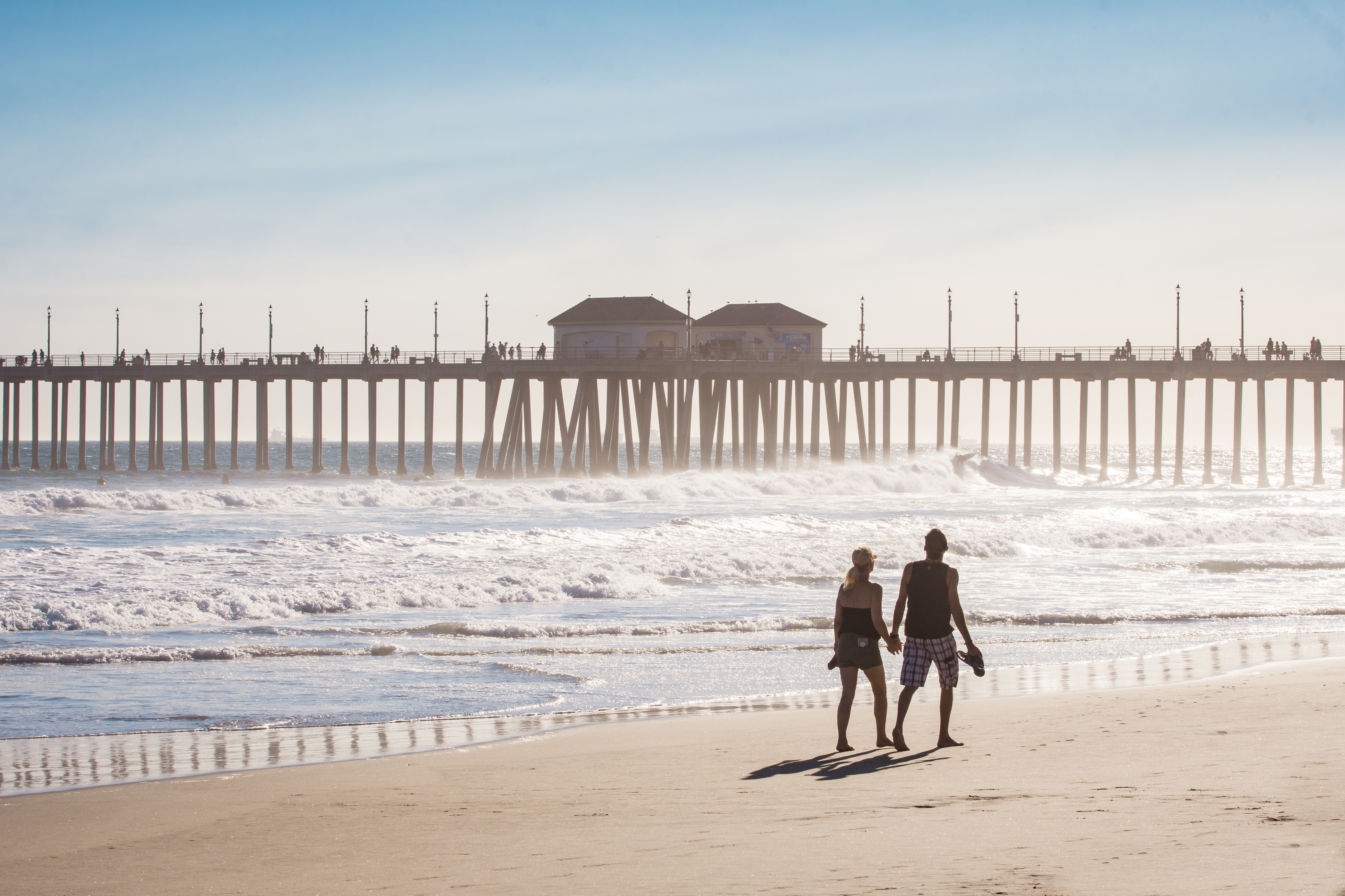 A couple walks along the sandy shores of Huntington Beach, California, hand in hand. Waves crash nearby as a long pier with people strolling on it stretches over the water beneath a clear blue sky.