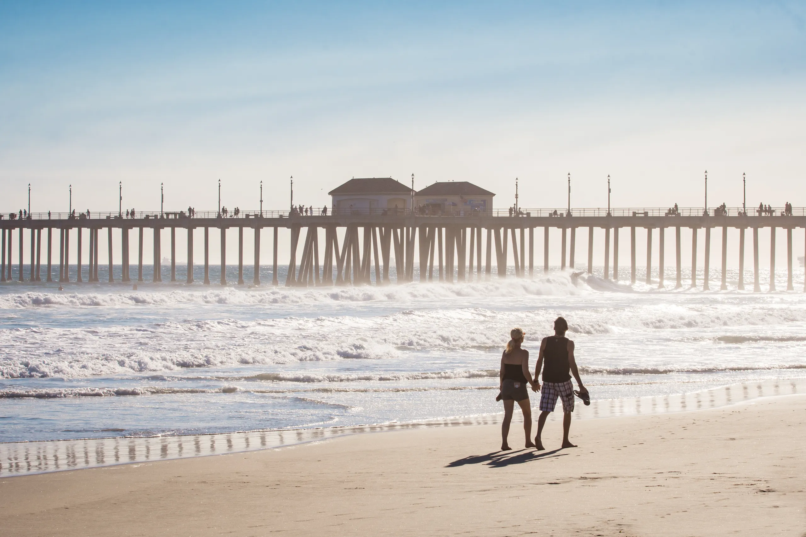 A couple walks along the sandy shores of Huntington Beach, California, hand in hand. Waves crash nearby as a long pier with people strolling on it stretches over the water beneath a clear blue sky.
