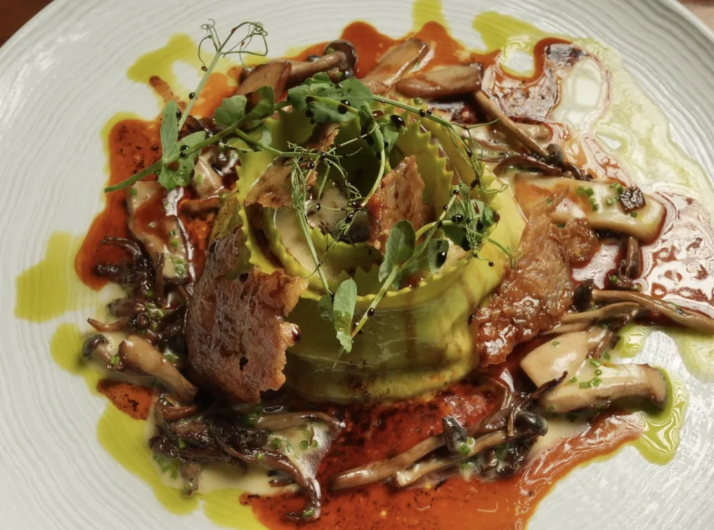 A gourmet dish featuring a green spiral of avocado topped with microgreens, surrounded by mushrooms, sauce, and drizzles of oil, presented on a white plate.