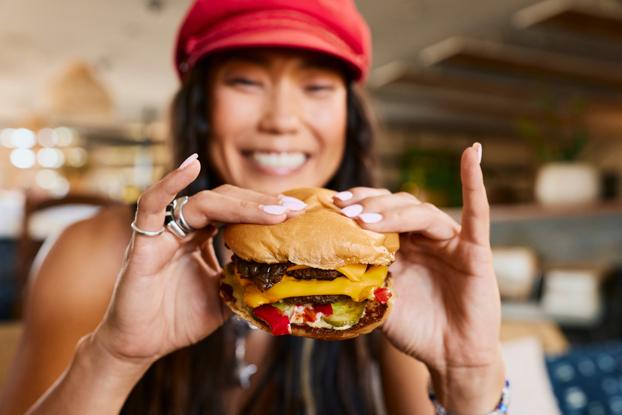 A person wearing a red cap and smiling holds a cheeseburger with lettuce, tomato, cheese, pickles, and a beef patty in both hands, ready to take a bite. The background is blurred.