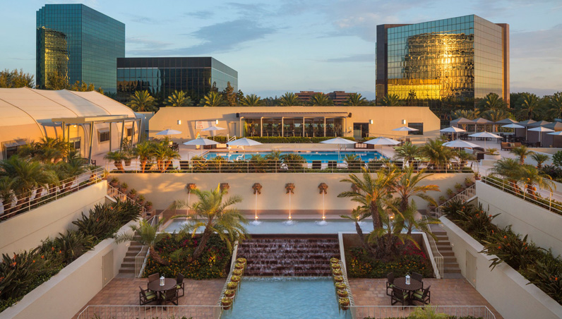 A modern hotel with a landscaped courtyard, cascading water features, palm trees, and an outdoor pool, surrounded by lounge chairs and umbrellas, with glass office buildings in the background.