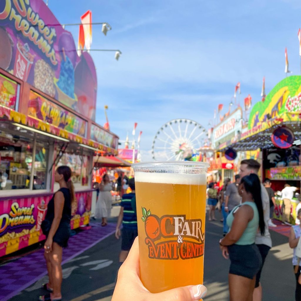 A hand holds a cup of beer with “OC Fair & Event Center” printed on it at a lively fair, with colorful food stalls, people walking, and a Ferris wheel in the background under a blue sky.
