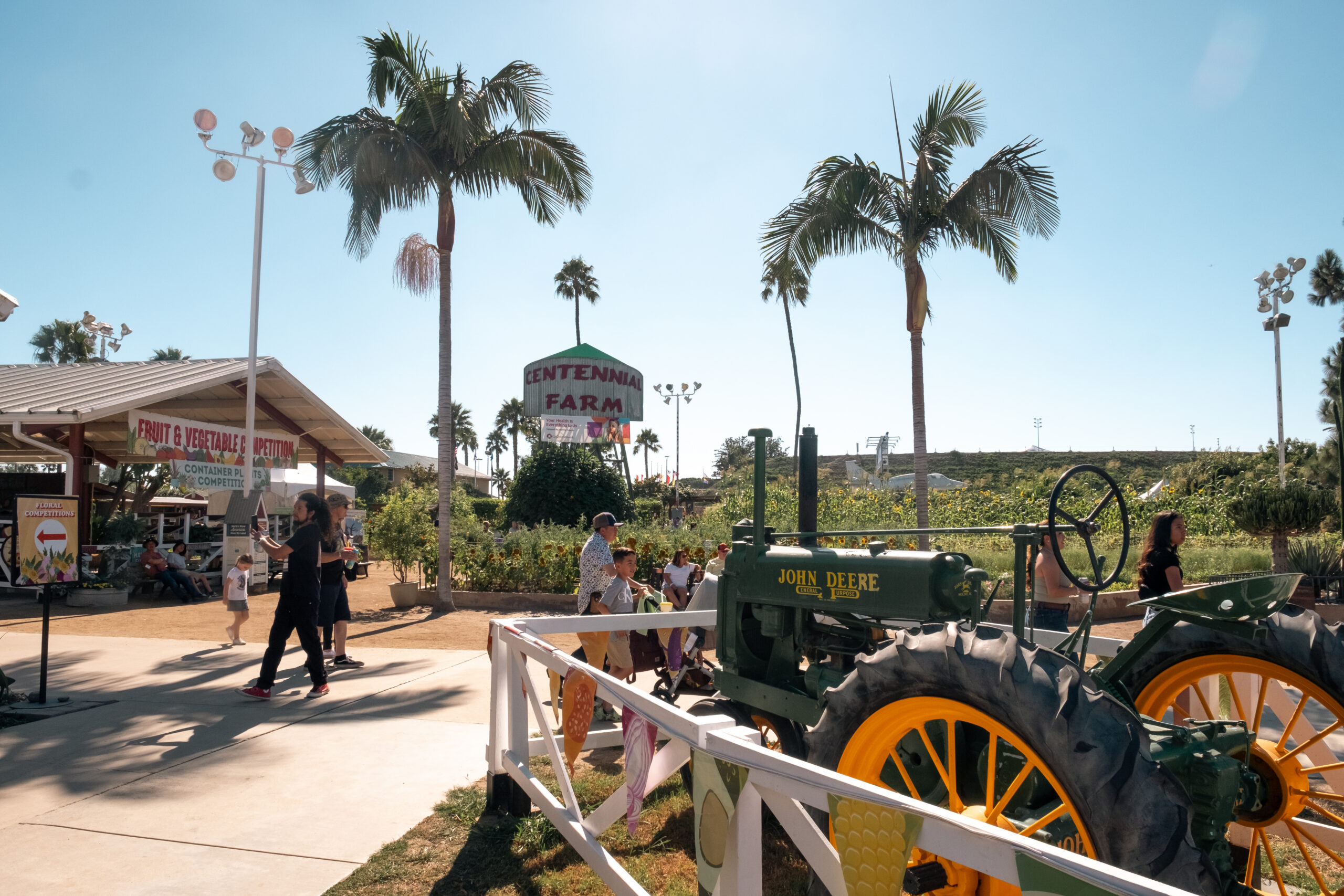 People walk near a vintage green John Deere tractor at Centennial Farm during the OC Fair Costa Mesa, surrounded by palm trees and garden beds. A fruit and vegetable stand is on the left, and the sky is bright and clear.