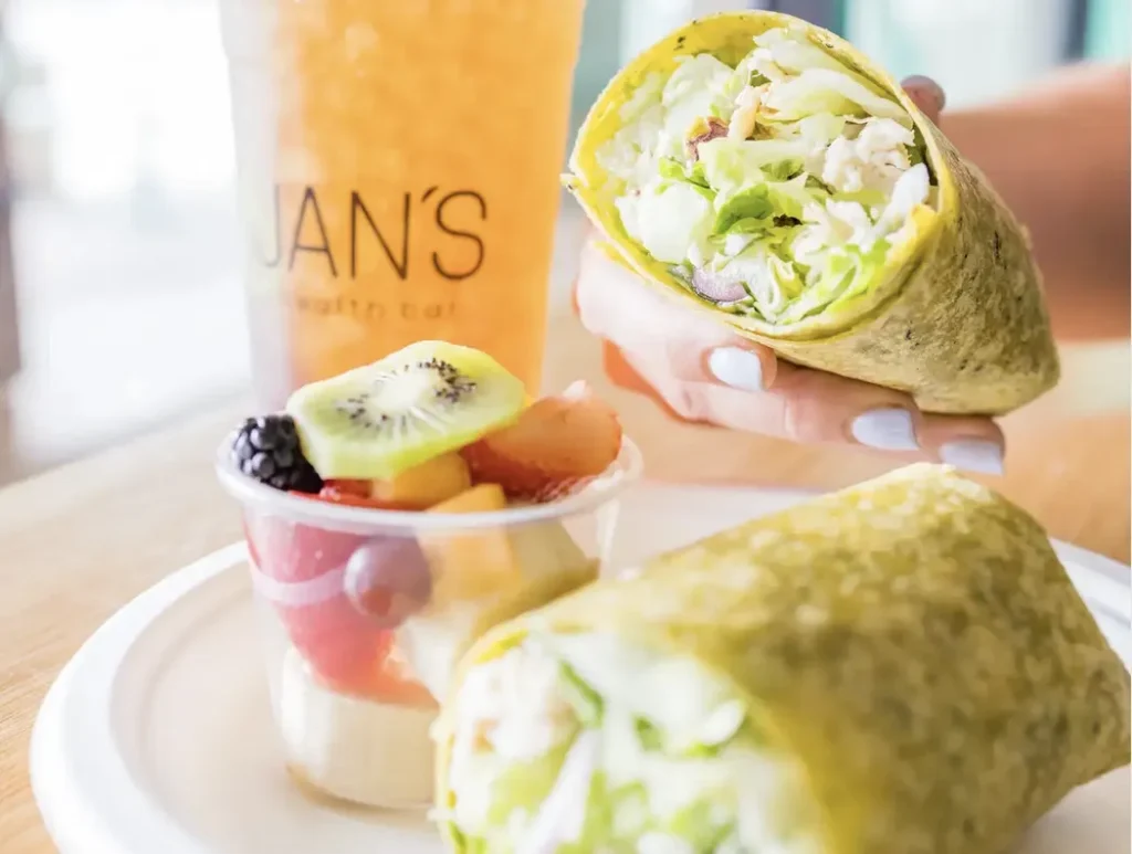 A hand holds half of a wrap filled with lettuce, chicken, and other veggies; the other half sits on a plate next to a cup of mixed fruit and a cold drink in a cup labeled JANS Health Bar.