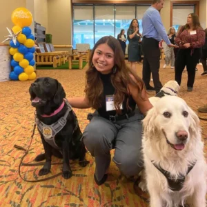 A woman kneels indoors between a black Labrador in a service vest and a white Golden Retriever. People and balloon decorations are visible in the background at what appears to be an event.