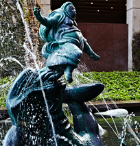 Bronze fountain sculpture of a joyful, plump child with arms outstretched, playfully riding on a fish with water spraying around them—an enchanting highlight of the Costa Mesa Art Walk’s outdoor setting.