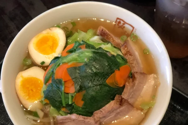 A bowl of Kitakata ramen topped with sliced pork belly, a soft-boiled egg, and greens, served with a side salad and a glass of iced tea at Ban Nai.