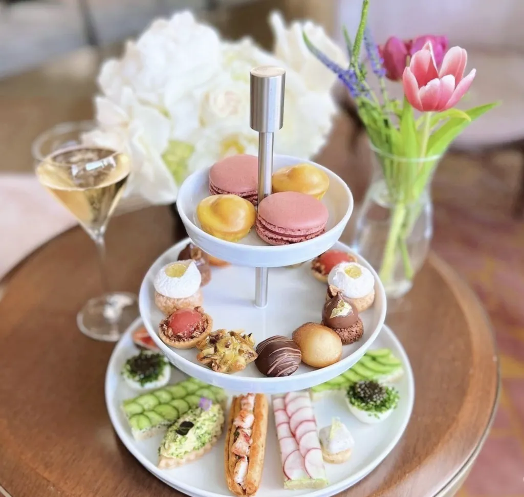 A two-tiered stand with assorted pastries, macarons, and savory snacks sits on a round table beside a glass of champagne and a vase with pink tulips and white flowers.