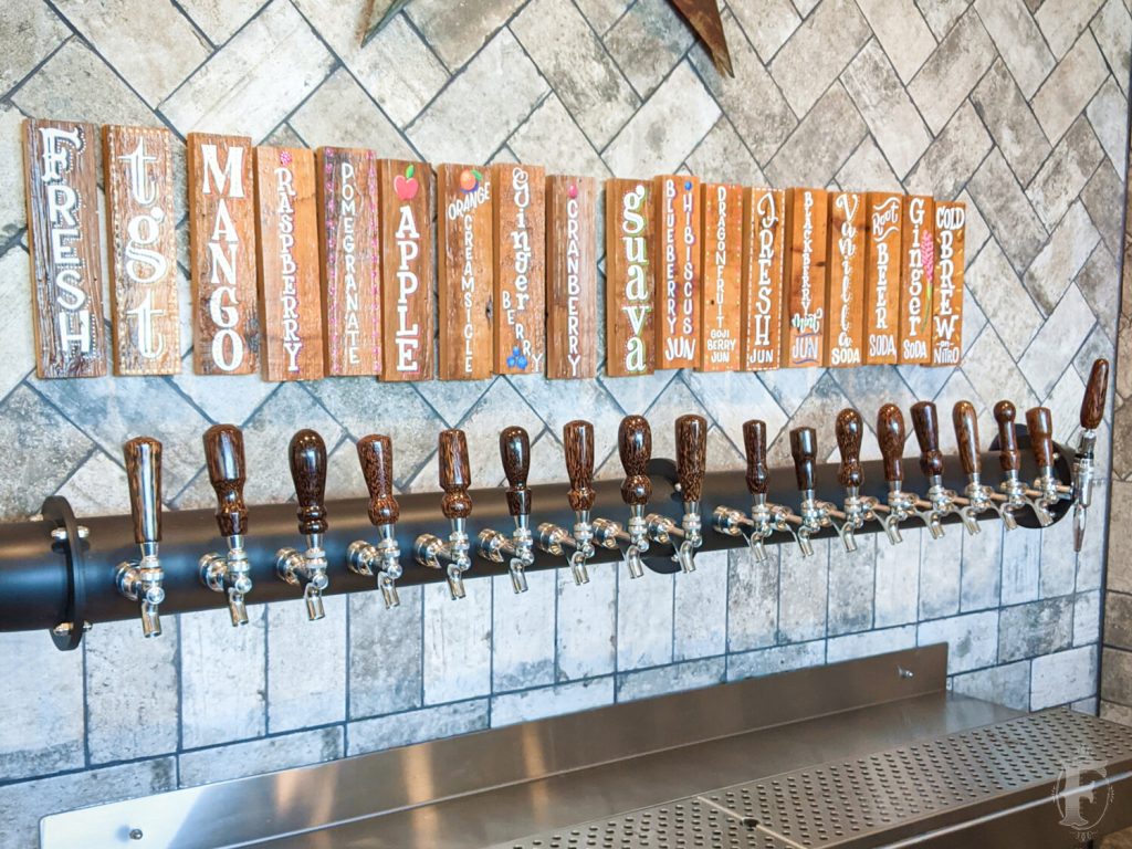 A row of beer taps is mounted on a tiled wall, each with a wooden label above indicating different drink flavors, such as mango, raspberry, apple, and others.