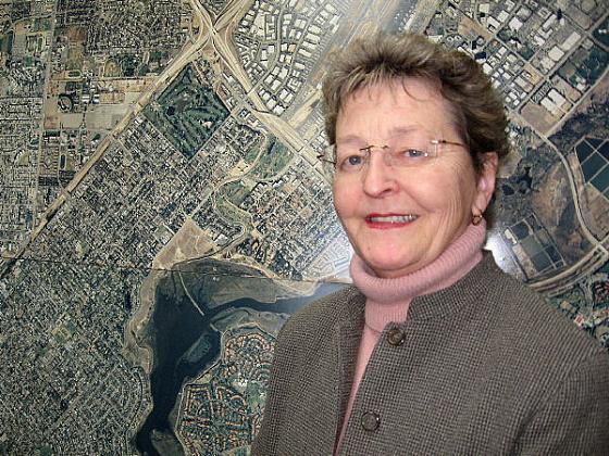 A woman with short hair, glasses, and a brown blazer over a pink turtleneck stands smiling in front of an aerial map of a city.