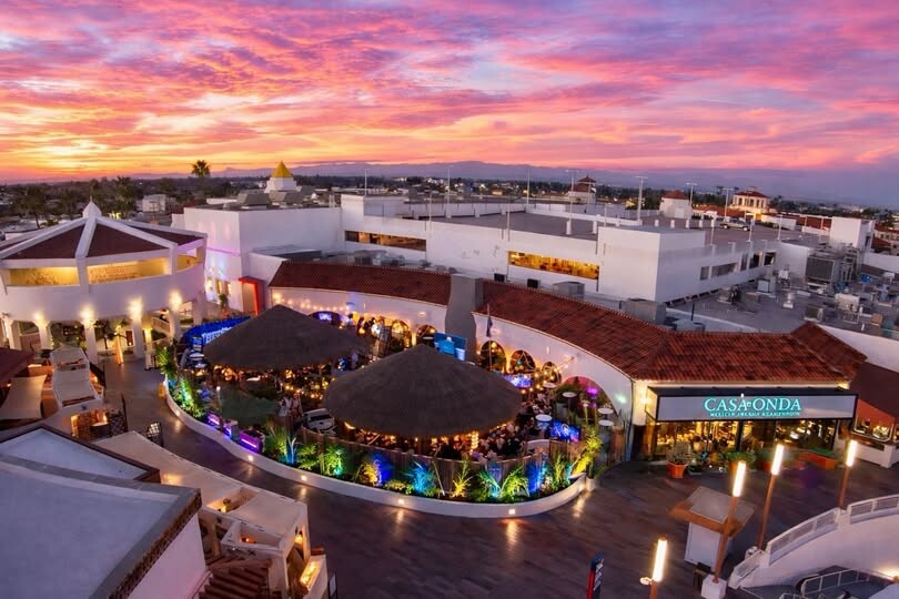 A vibrant outdoor plaza at sunset features white buildings, colorful lights, and thatched-roof dining areas. The sky is painted with dramatic pink and orange clouds. Signs read “CASA ONDA.”.