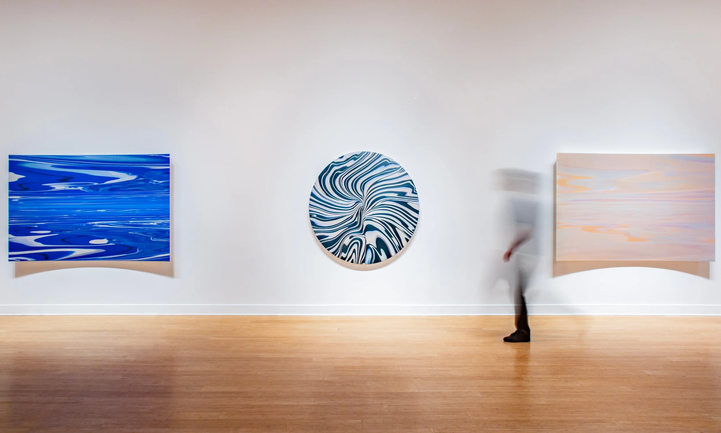 A person walks past three abstract paintings on a white gallery wall: a blue rectangular piece, a circular marbled piece, and a pastel rectangular piece. The person appears blurred in motion.