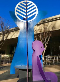 A colorful outdoor sculpture features a blue abstract figure with uplifted arms, a circular striped design above, and a seated purple human silhouette in front, set against a building and clear blue sky.