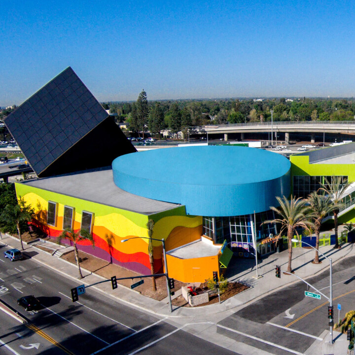 Aerial view of a colorful modern building with a large tilted black cube on its roof, a blue circular section, and wavy rainbow-colored walls, located at a street intersection with palm trees and traffic lights.