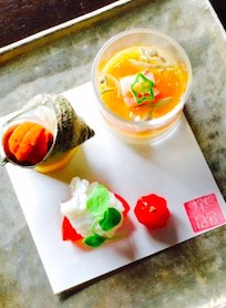 A Japanese appetizer plate at Hana Re Sushi Costa Mesa features a sea snail shell with orange uni, a glass cup with yellow and green garnish, and two pieces of sashimi with green leaf accents on a white square plate.