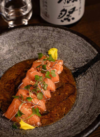 Slices of salmon sashimi topped with microgreens and sesame seeds, served on a rustic stone plate with brown sauce and a yellow flower garnish at Yuzu Sushi Costa Mesa. A glass and a sake bottle are in the background.