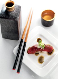A piece of sushi with sauce is served on a white square plate beside chopsticks, a black sake bottle, and a small cup with amber liquid at Hamamori Costa Mesa.