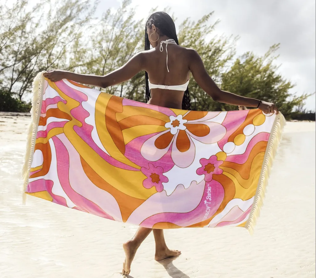 A woman in a white bikini stands on a sandy beach, holding a colorful towel with bold, retro-style floral patterns in pink, orange, yellow, and white. Trees and clear sky are visible in the background.