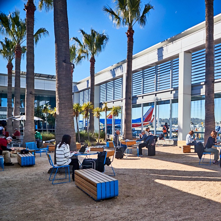 People sit on benches and chairs under palm trees in an airport’s outdoor seating area. Reflections of a Southwest Airlines plane and blue sky appear in the glass windows, with travelers discussing getting to Costa Mesa from the airport.