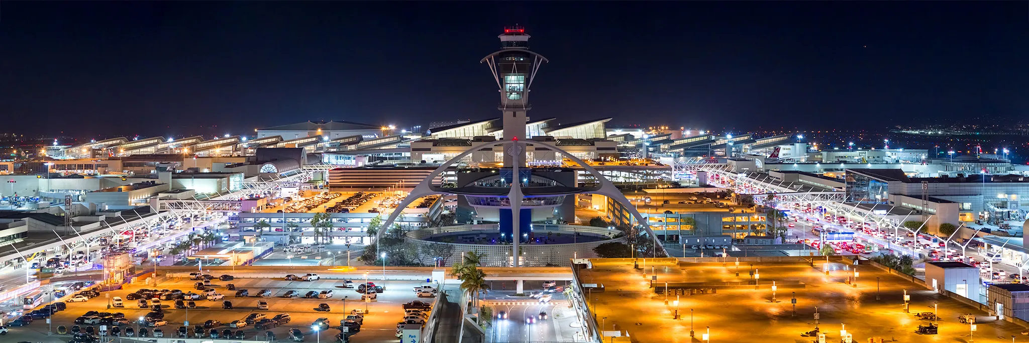 Night view of a large, brightly lit airport with a central control tower and the iconic Theme Building, where glowing streetlights guide travelers getting to Costa Mesa from airport terminals surrounded by parked cars.
