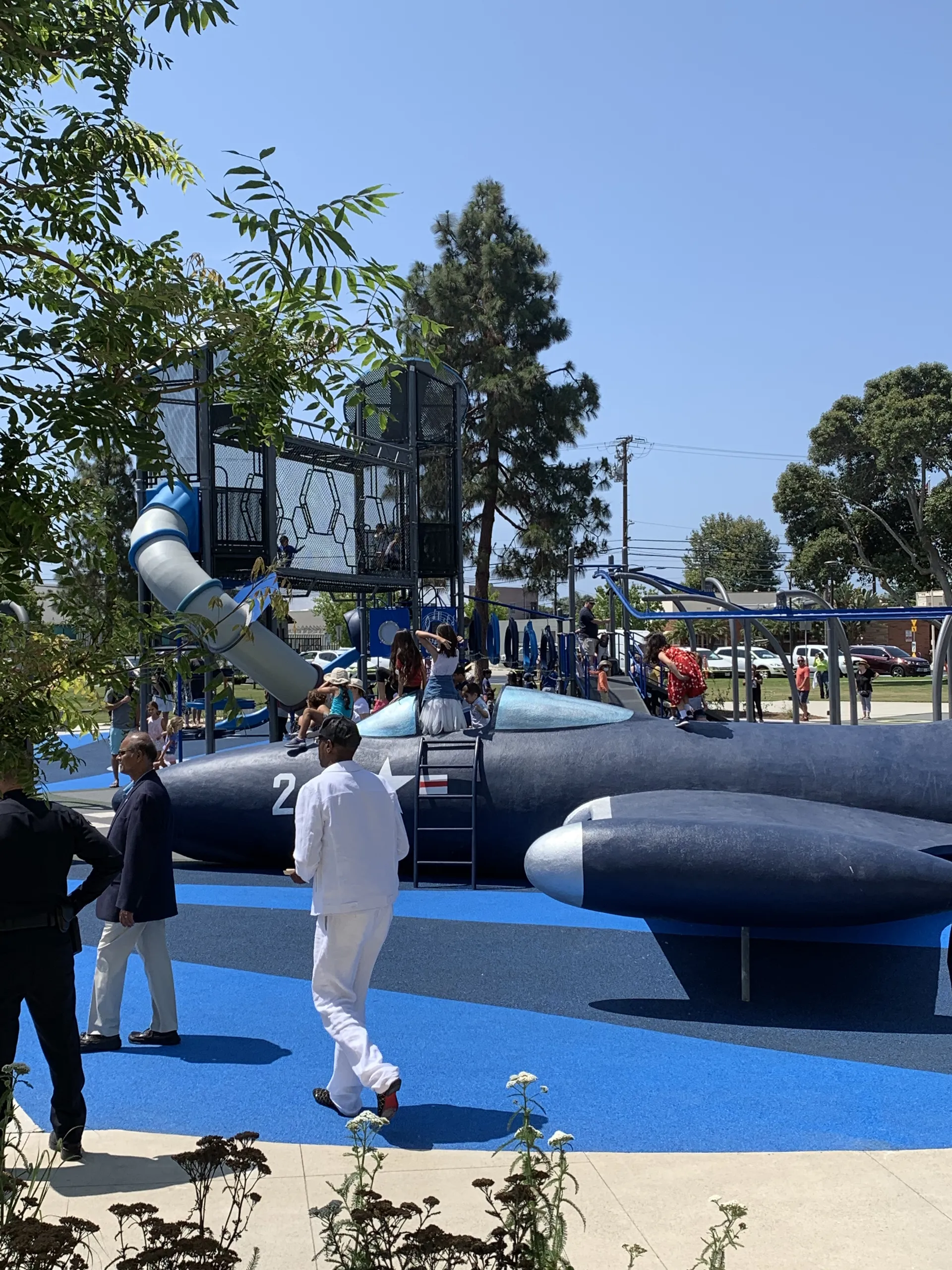Children play on a blue airplane-themed playground structure with a slide at Lions Park Costa Mesa, surrounded by adults and trees on a sunny day. Some children are climbing while others are inside the plane.