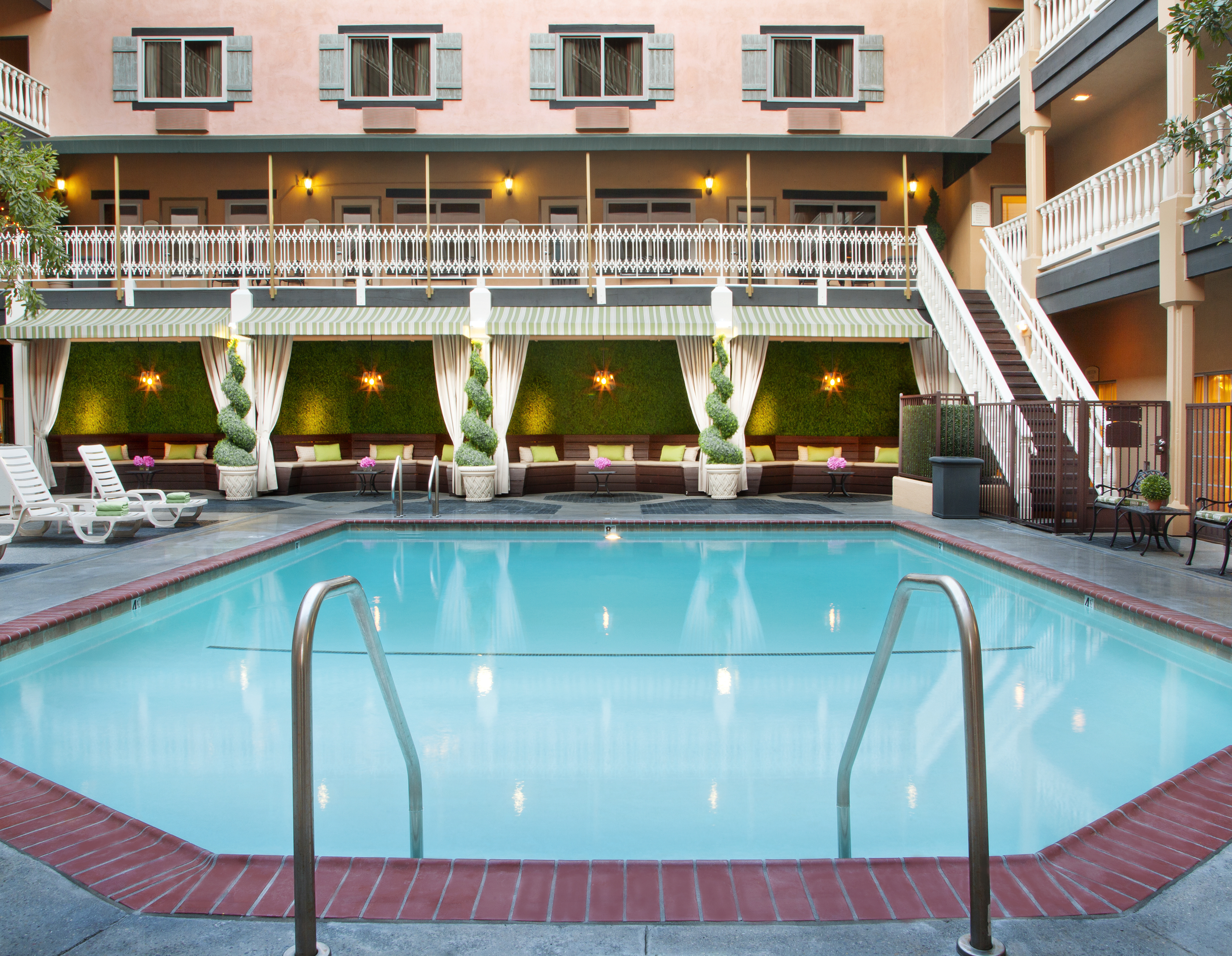 An outdoor hotel pool with clear water at Ayres Hotels, surrounded by lounge chairs, greenery, and cabanas. The building features balconies and large windows, creating a relaxing, inviting atmosphere.