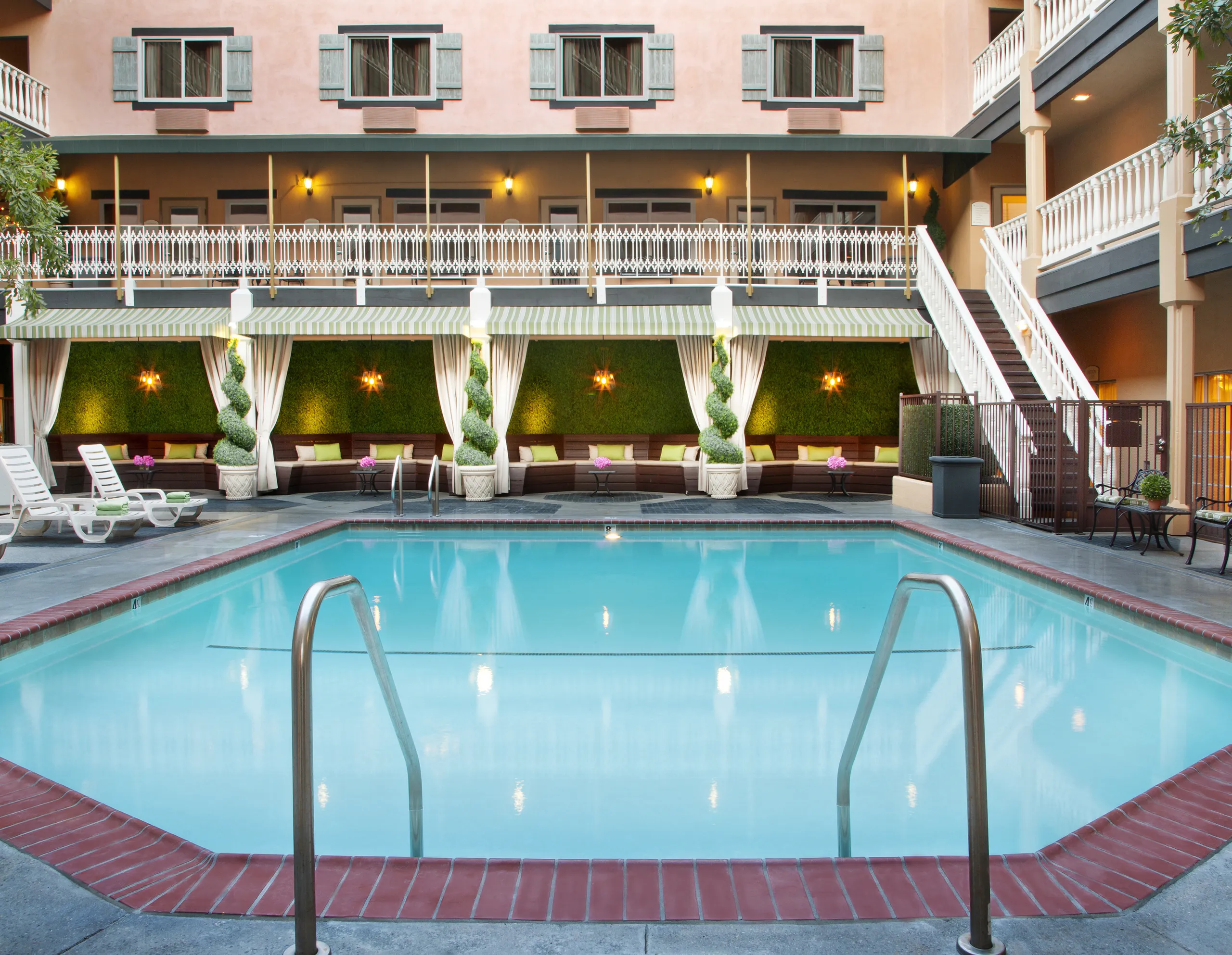 An outdoor hotel pool with clear water at Ayres Hotels, surrounded by lounge chairs, greenery, and cabanas. The building features balconies and large windows, creating a relaxing, inviting atmosphere.