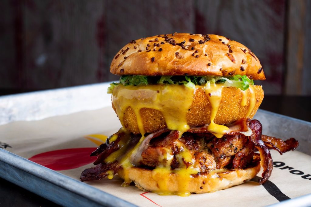 A stacked burger with bacon, grilled chicken, lettuce, a large fried onion ring, and melted cheese on a seeded bun, served on a metal tray with parchment paper.