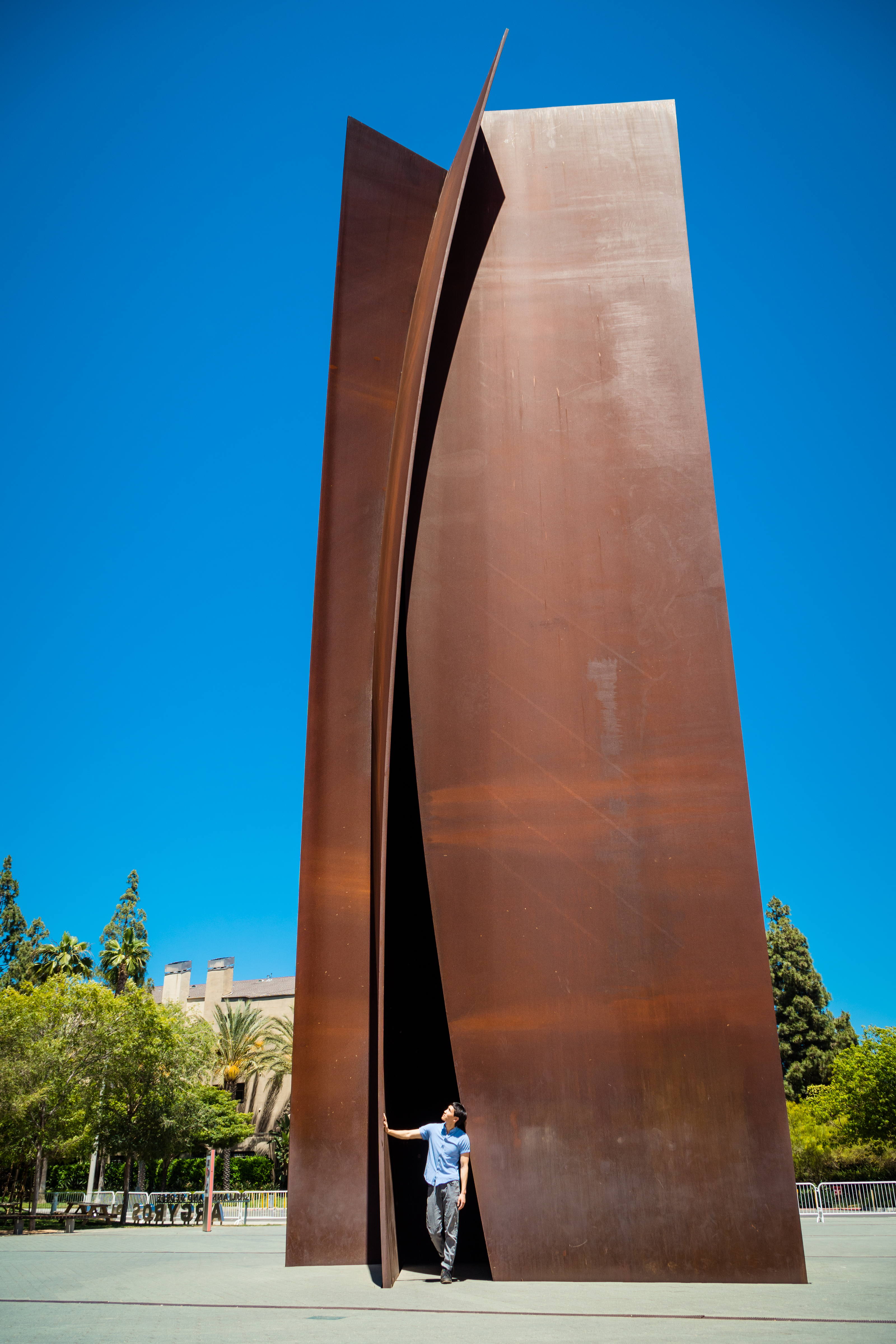 A person stands at the base of the tall, abstract rust-colored Connector sculpture in Costa Mesa, reaching up with one hand. The sky is clear and blue, with trees and a building visible in the background.