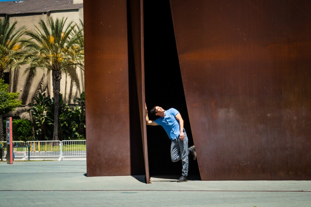 A person in a blue shirt and jeans leans playfully against the edge of the large Connector sculpture outdoors in Costa Mesa, with palm trees and a building in the background.