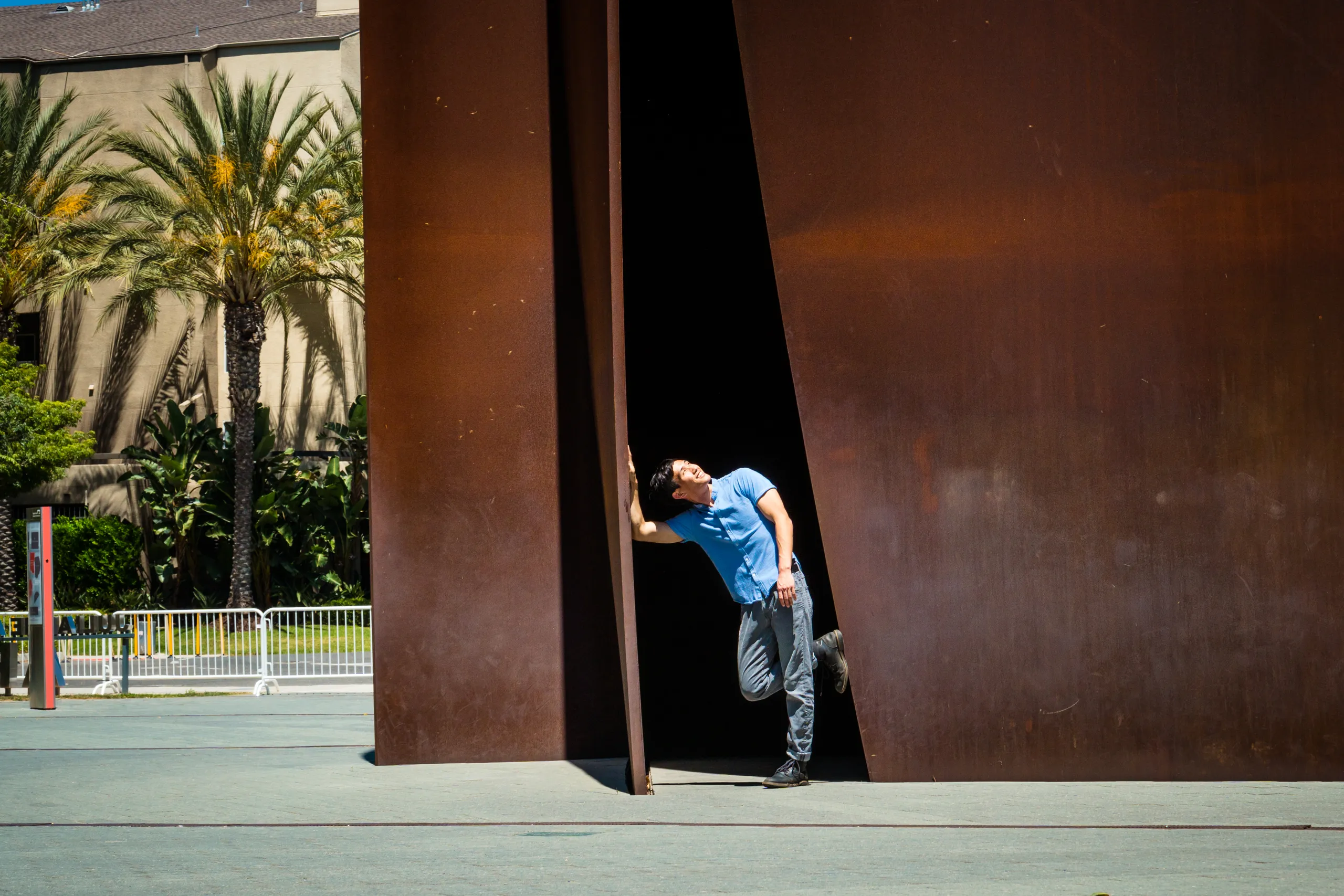 A person in a blue shirt and jeans leans playfully against the edge of the large Connector sculpture outdoors in Costa Mesa, with palm trees and a building in the background.