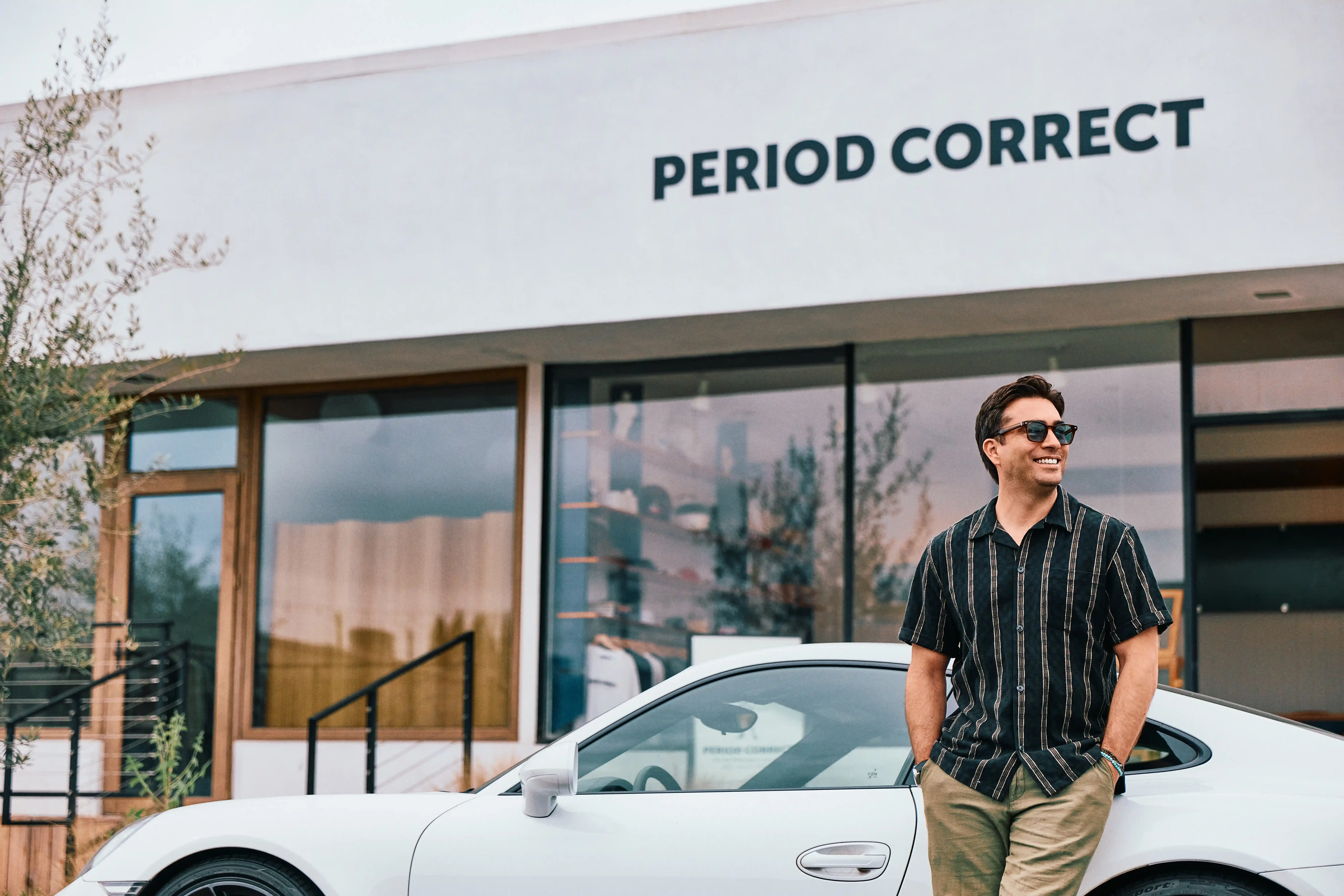 A man wearing sunglasses and a striped shirt leans against a white car parked in front of a modern building with large windows and a sign that reads PERIOD CORRECT.