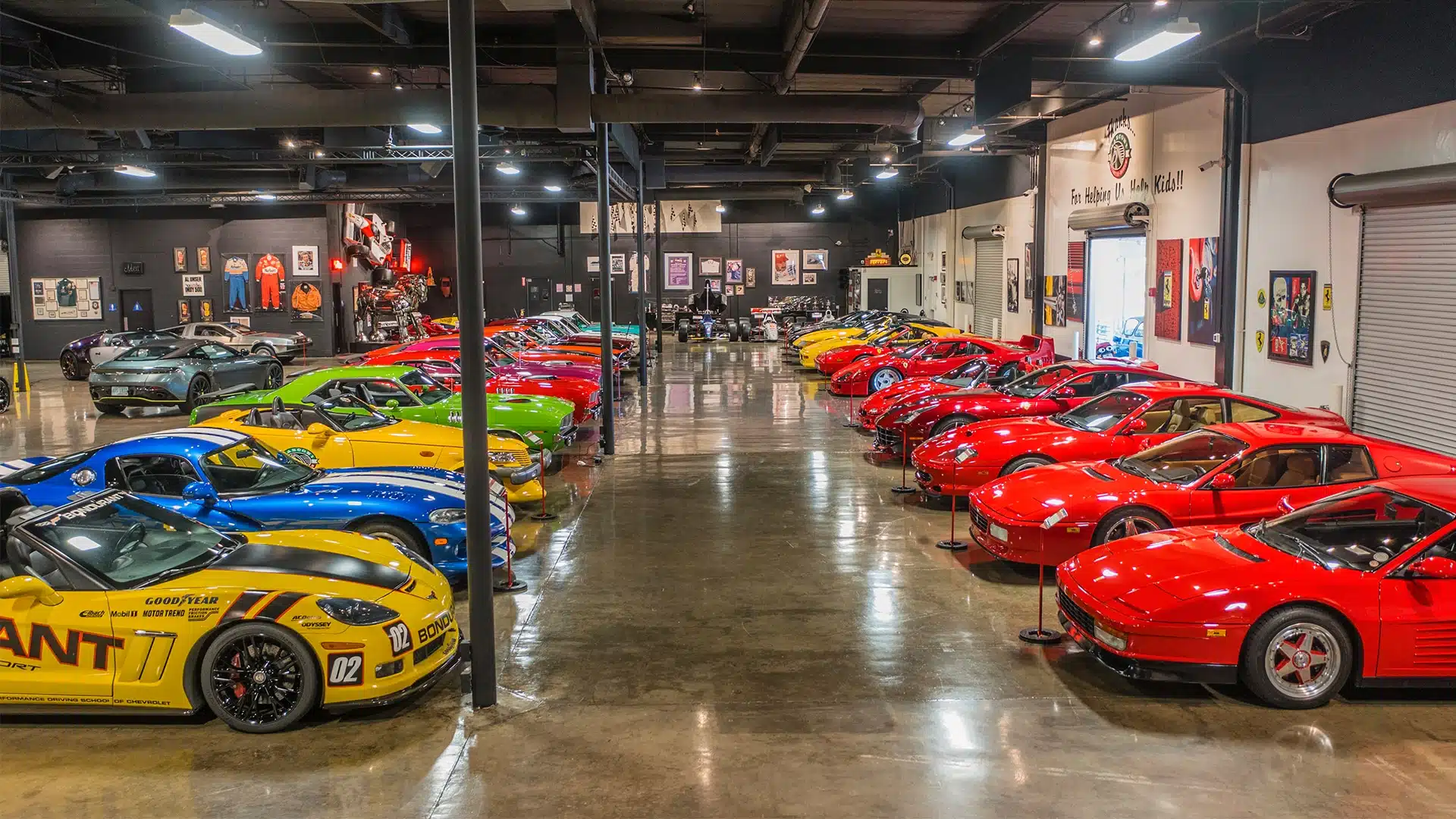 A spacious showroom features two rows of brightly colored sports cars, including red, yellow, blue, and green vehicles, displayed under bright lights with automotive memorabilia on the walls.