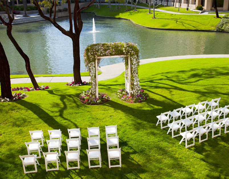 Rows of white chairs are arranged on a green lawn at Marriott Costa Mesa, facing a flower-covered wedding arch, with a pond and fountain in the background under sunny skies.
