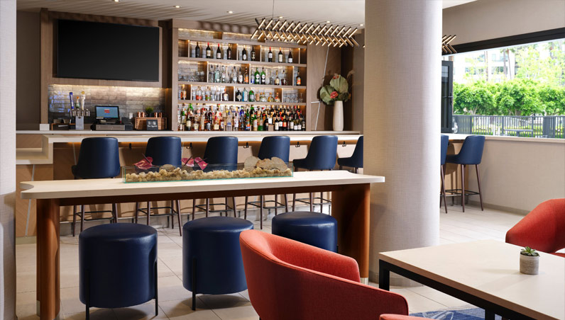 Modern hotel bar at Marriott Costa Mesa with blue stools at the counter, red chairs around tables, shelves stocked with bottles, a large TV above the bar, and large windows letting in natural light.