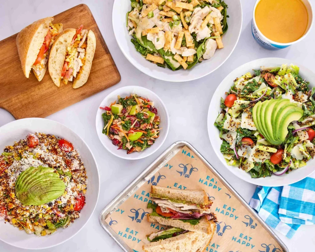 A colorful spread of salads, sandwiches, and soup, including a chicken salad, avocado salads, fresh sandwiches, a bowl of soup, and a fruit salad, all arranged on a white surface with a blue checkered napkin.