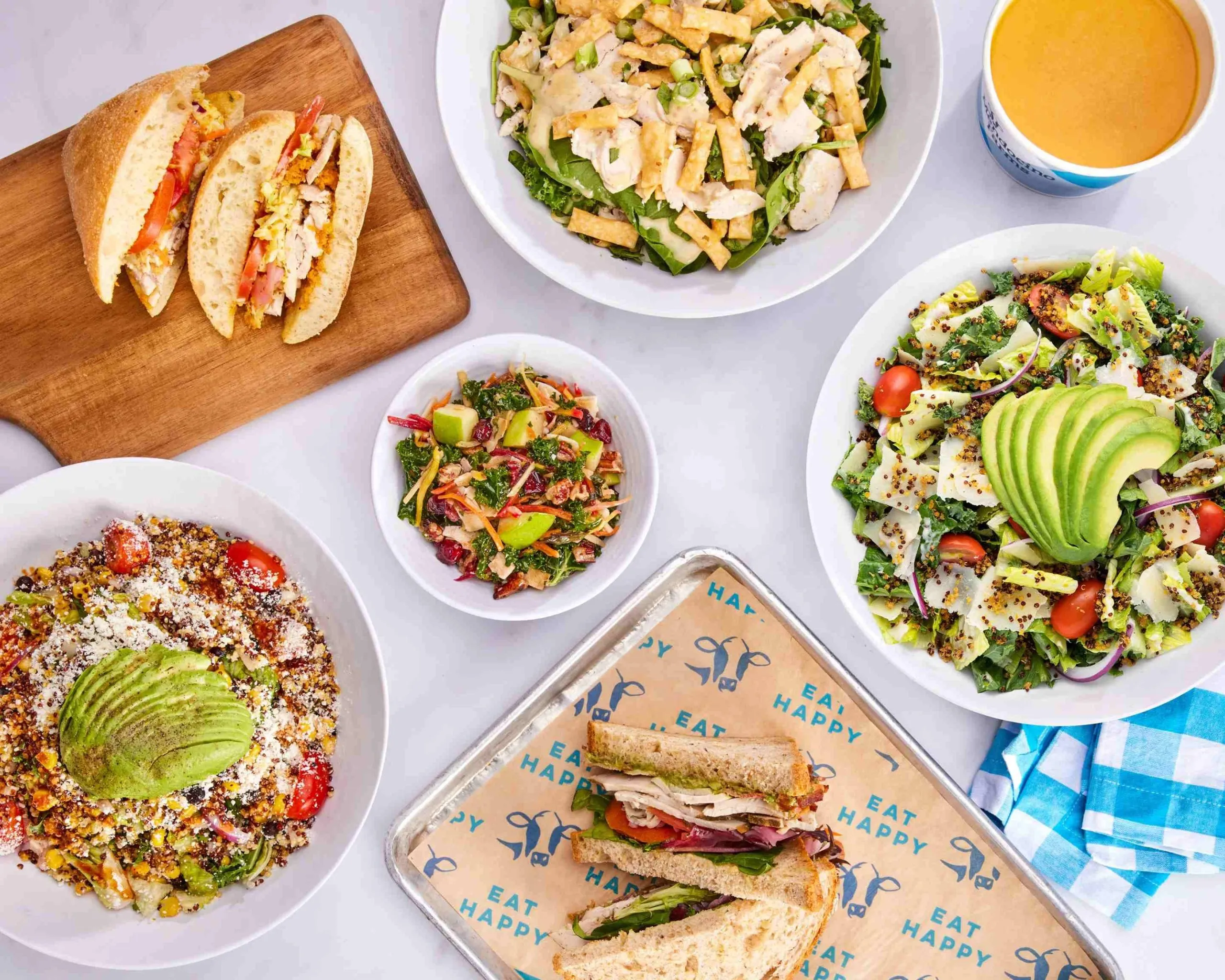 A colorful spread of salads, sandwiches, and soup, including a chicken salad, avocado salads, fresh sandwiches, a bowl of soup, and a fruit salad, all arranged on a white surface with a blue checkered napkin.