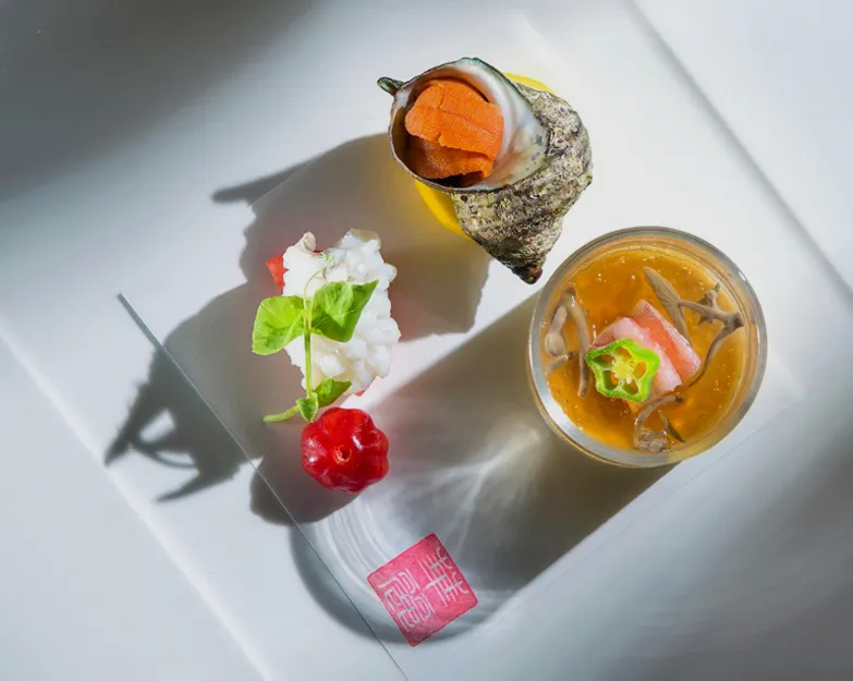 A gourmet Japanese dish features a sea snail shell with orange seafood, a glass with soup and vegetables, a white seafood piece with a leaf garnish, and a small red garnish on a white square plate.