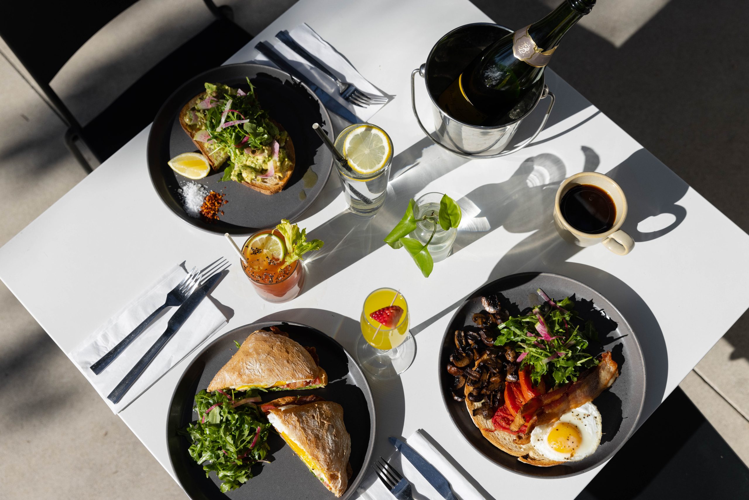 Overhead view of a table set for two with plates of sandwiches, salad, breakfast food, and drinks including coffee, sparkling wine, and cocktails, with utensils and napkins neatly arranged.