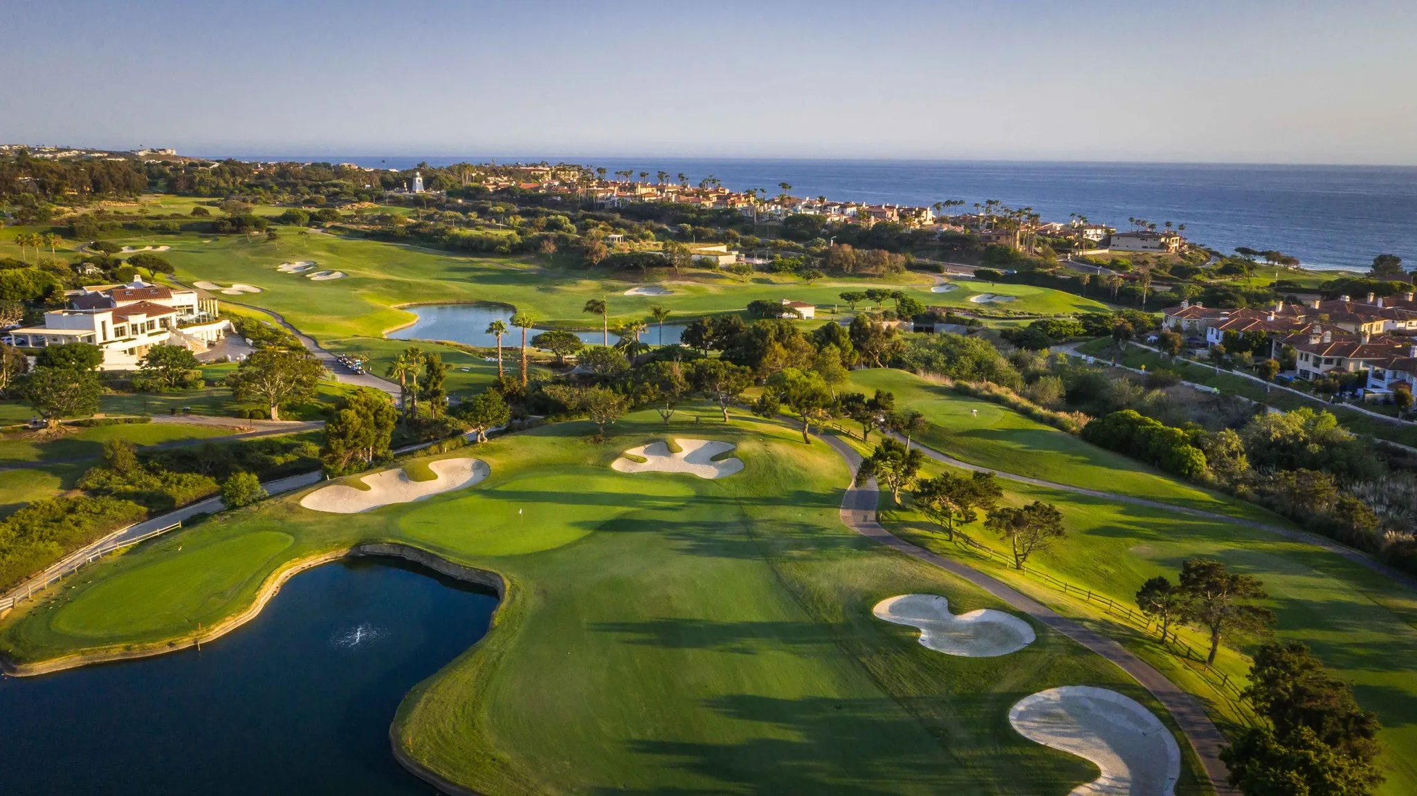 Aerial view of a lush, coastal golf course with well-manicured greens, sand bunkers, water features, nearby houses, and the ocean in the background under a clear sky.