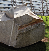 A large, rough-hewn stone sculpture with a metal plate embedded in its top stands on grass along the Costa Mesa Art Walk, framed by leafless trees and a modern building in the background.