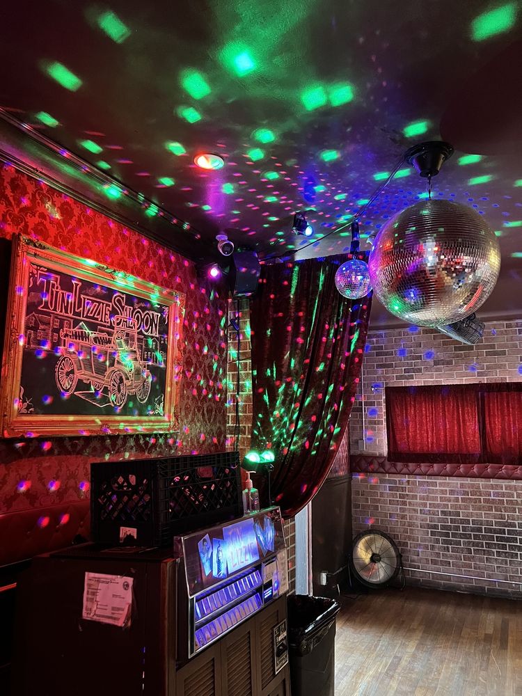 A disco ball and colored lights create vibrant reflections in a retro-style room with red curtains, brick walls, a jukebox, a gong, and a framed Tin Lizzie Saloon sign.