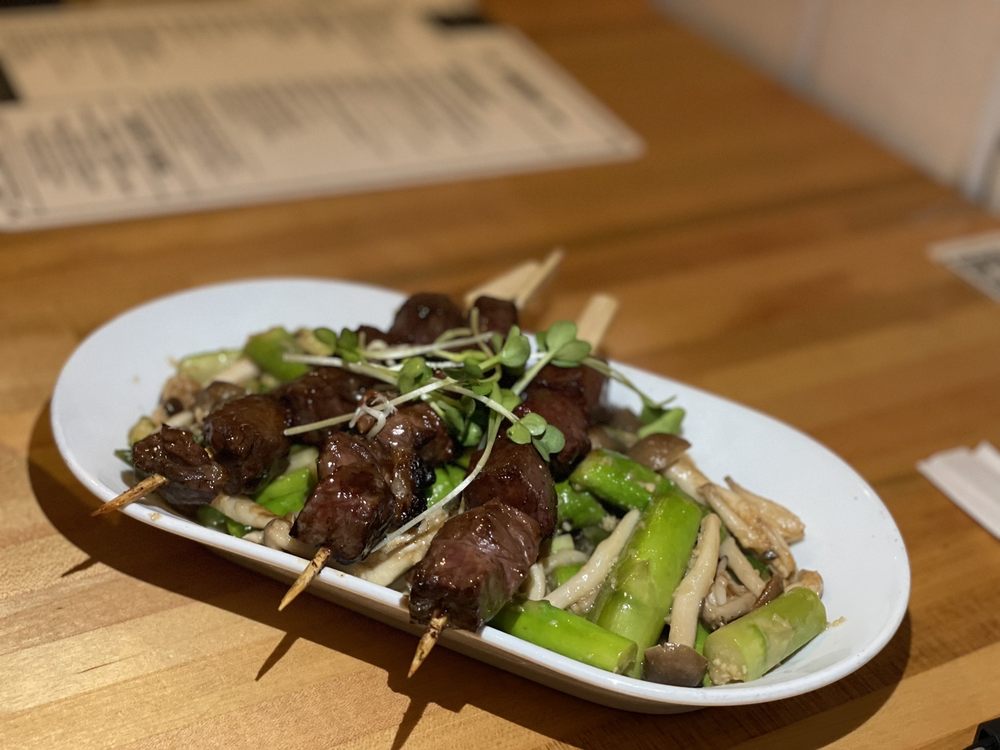A white oval plate with grilled beef skewers, asparagus, mushrooms, and garnished with microgreens, placed on a wooden table with a menu in the background.